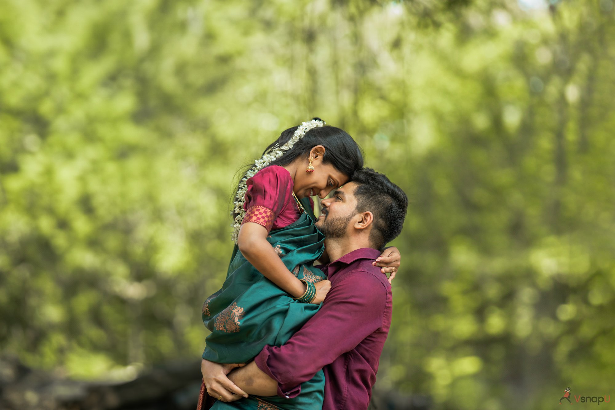 Pre-wedding shoot capturing an intimate moment of the groom lifting the bride in a saree, with vibrant greenery around them.